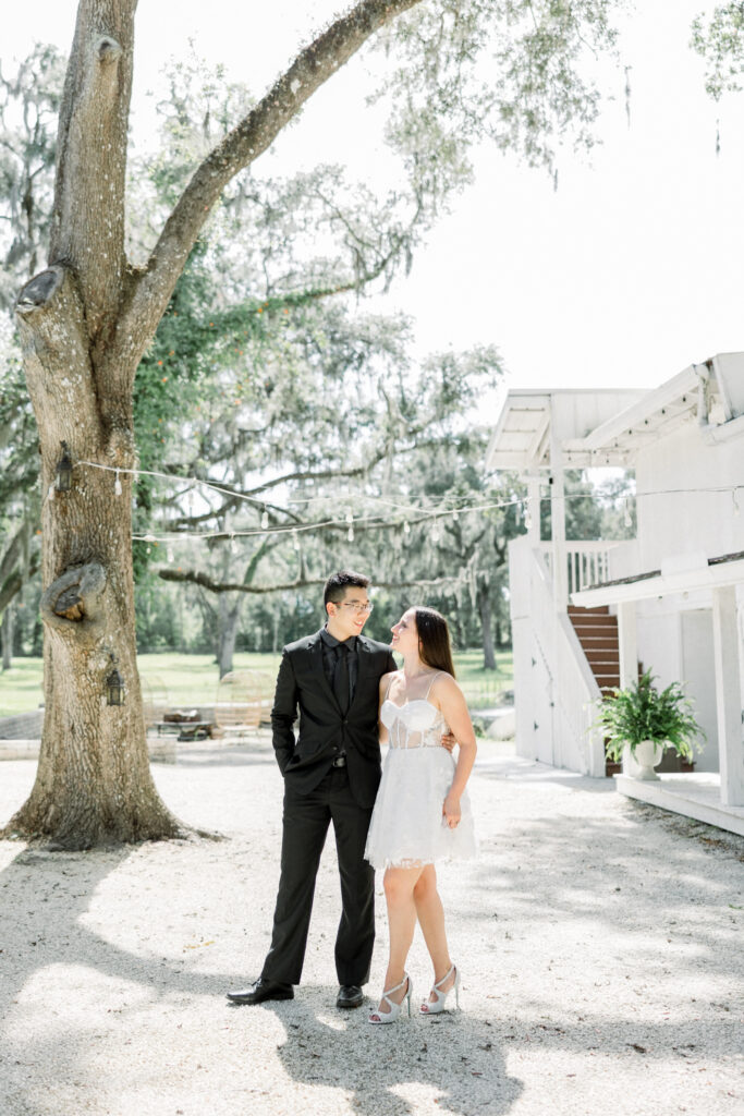 Wide shot of couple embracing in open field with Tuscan-inspired architecture in the background