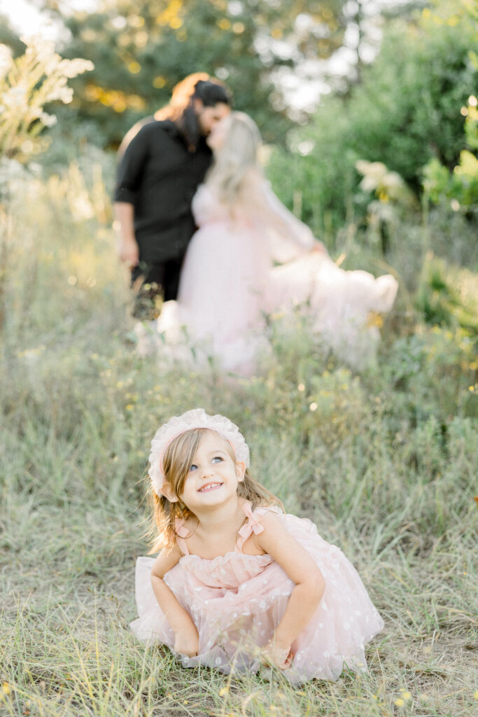 Toddler smiling with parents kissing behind